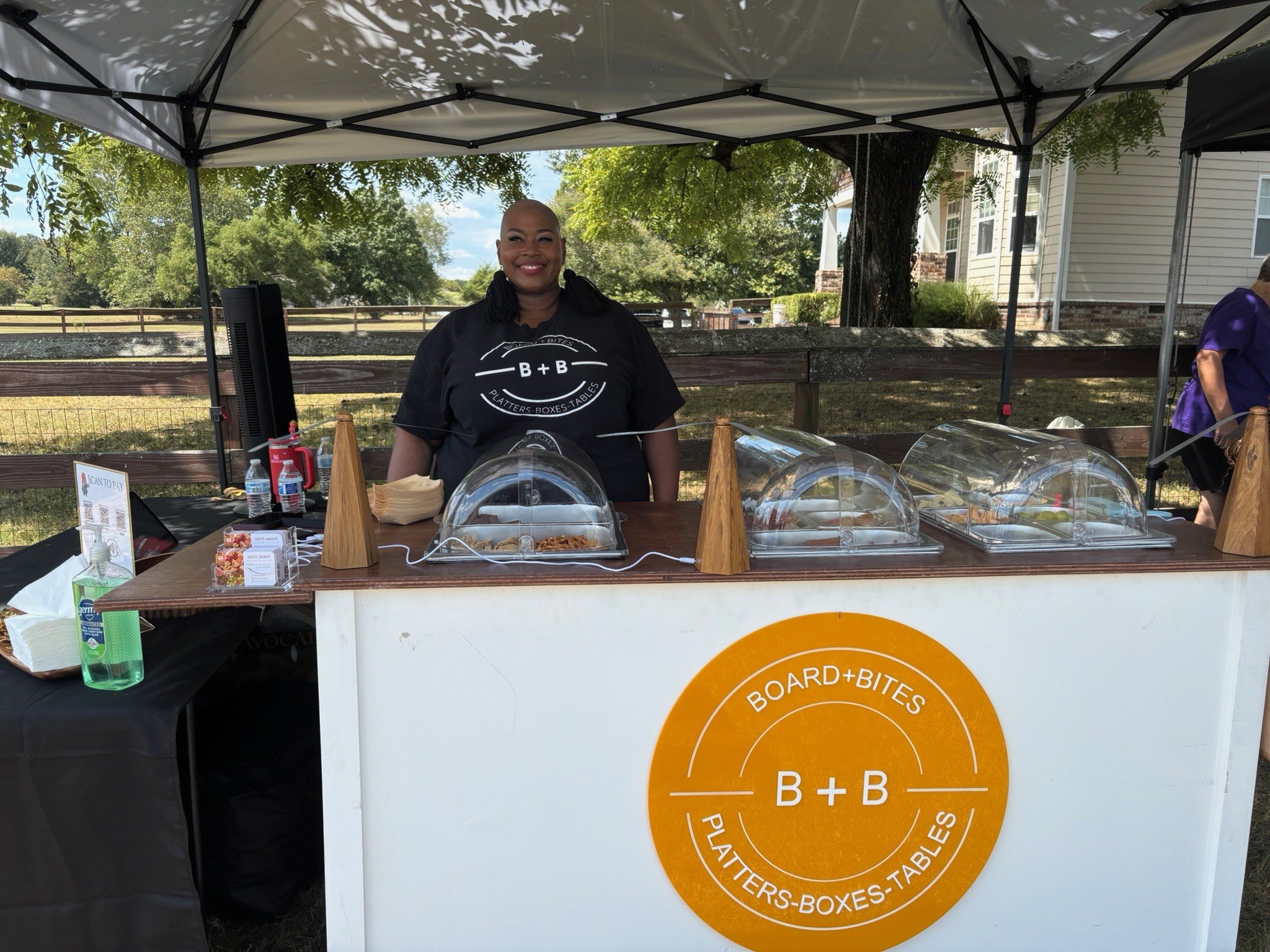 A woman standing behind a food stand under a canopy, smiling, with a sign that reads 'B + B' and mentions 'Platers, Boxes, Tables'; the stand has food covered with transparent domes and is set outdoors with trees and a house in the background.