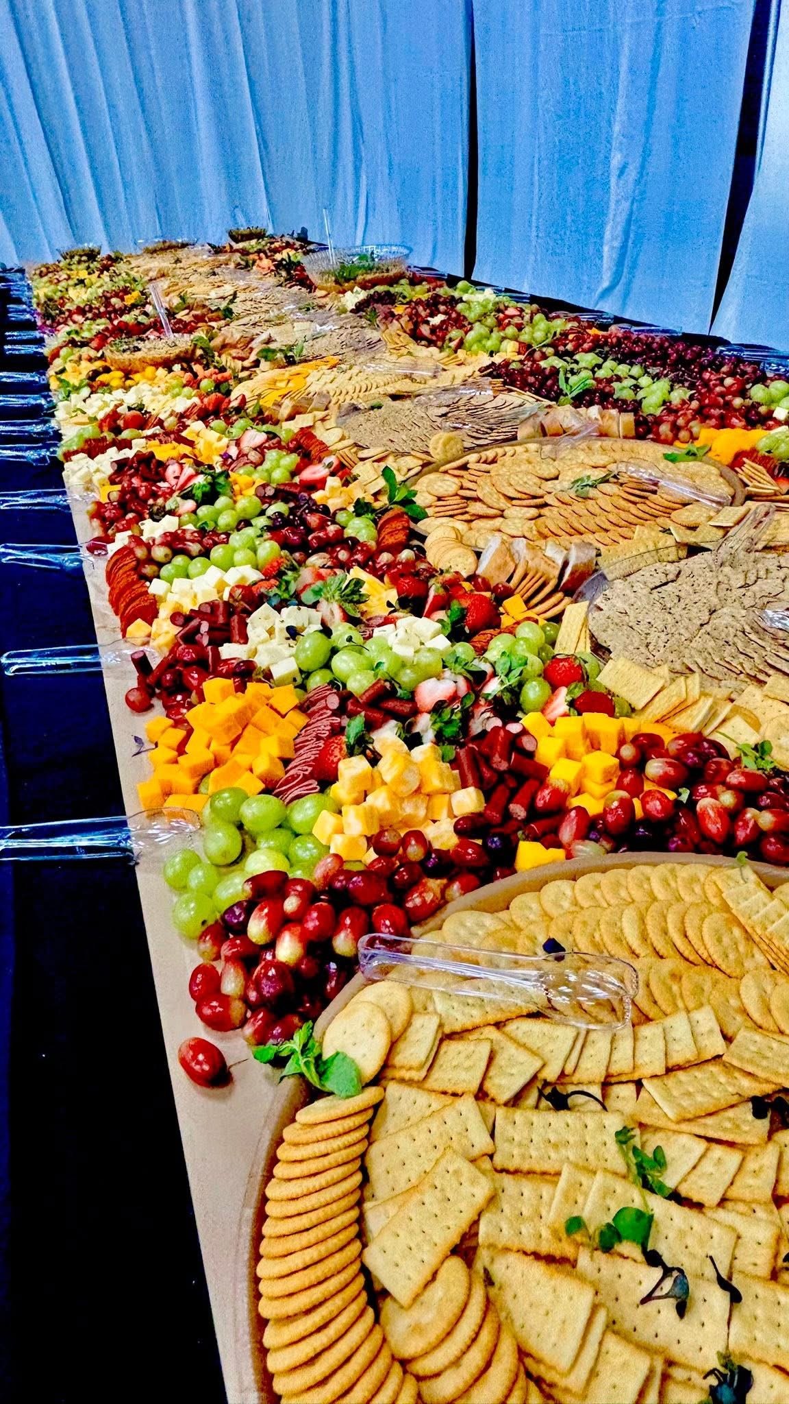 A long table filled with a variety of cheeses, crackers, grapes, and other fruits, set against a blue background.