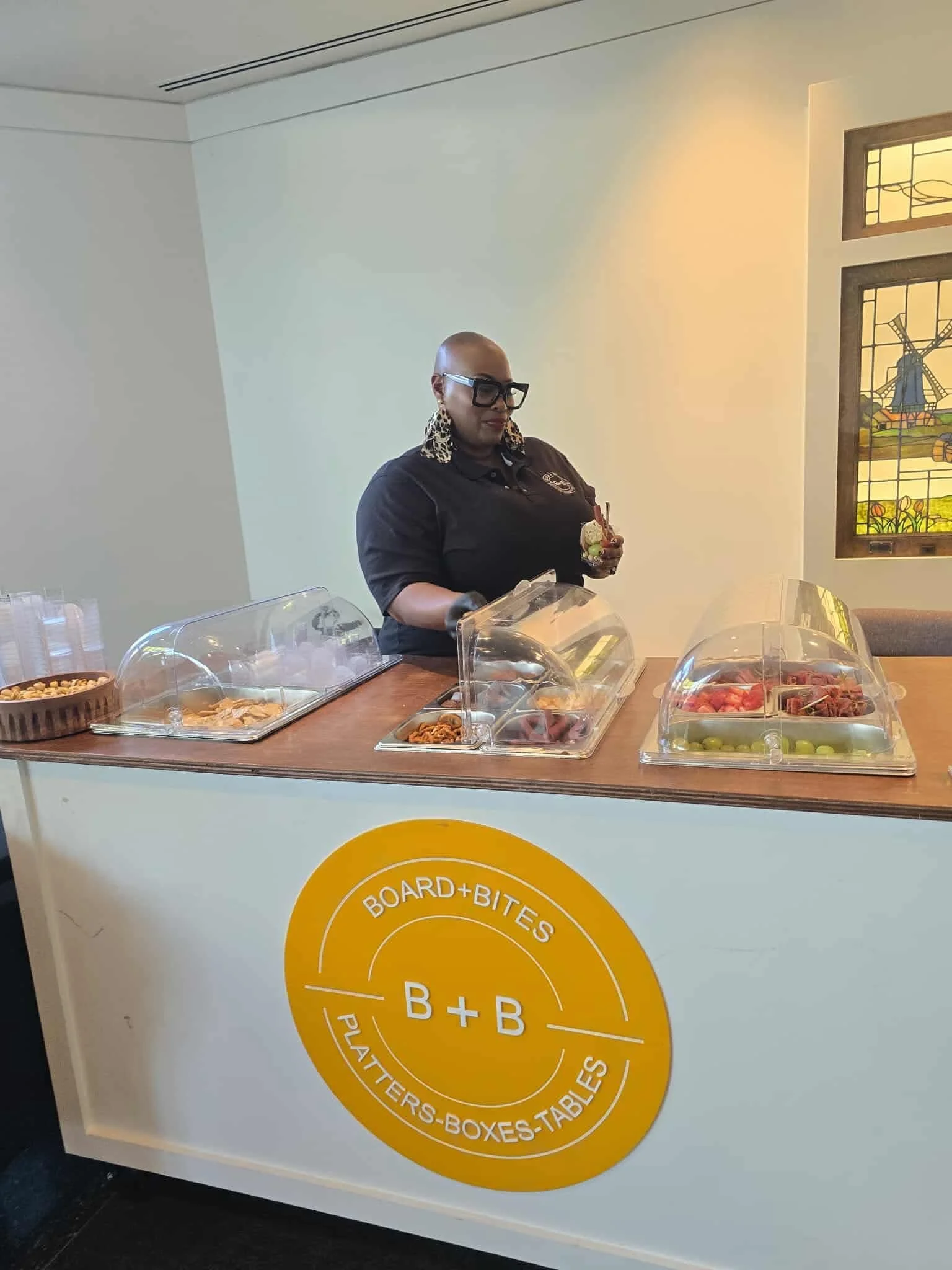 A woman serving food at a counter with a sign that reads "B+B Boards & Bites Platers-Boxes-Tables." She is wearing glasses, leopard print earrings, and a black shirt. There are food containers with different types of food on the counter.