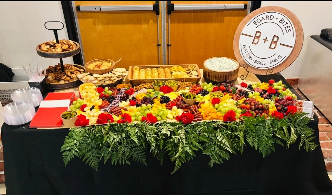 A table with a black tablecloth decorated with green fern leaves, filled with an assortment of cheeses, fruits, and snacks. There are bowls of green olives, a platter of crackers, grapes, and berries, and a three-tiered stand with nuts and cookies. A