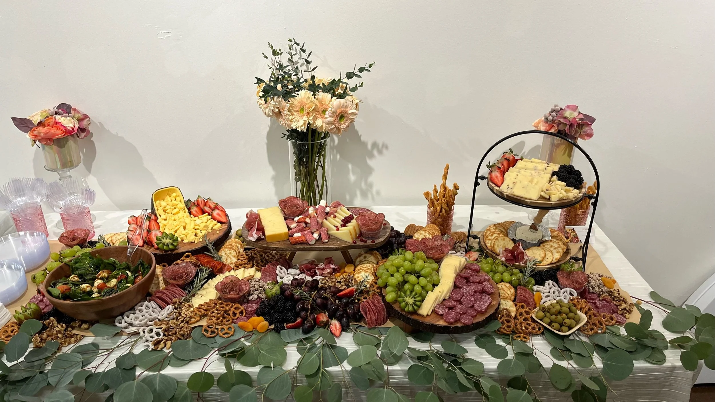 A table with an assortment of cheeses, cured meats, fruits, crackers, and nuts, decorated with flowers and green leaves.
