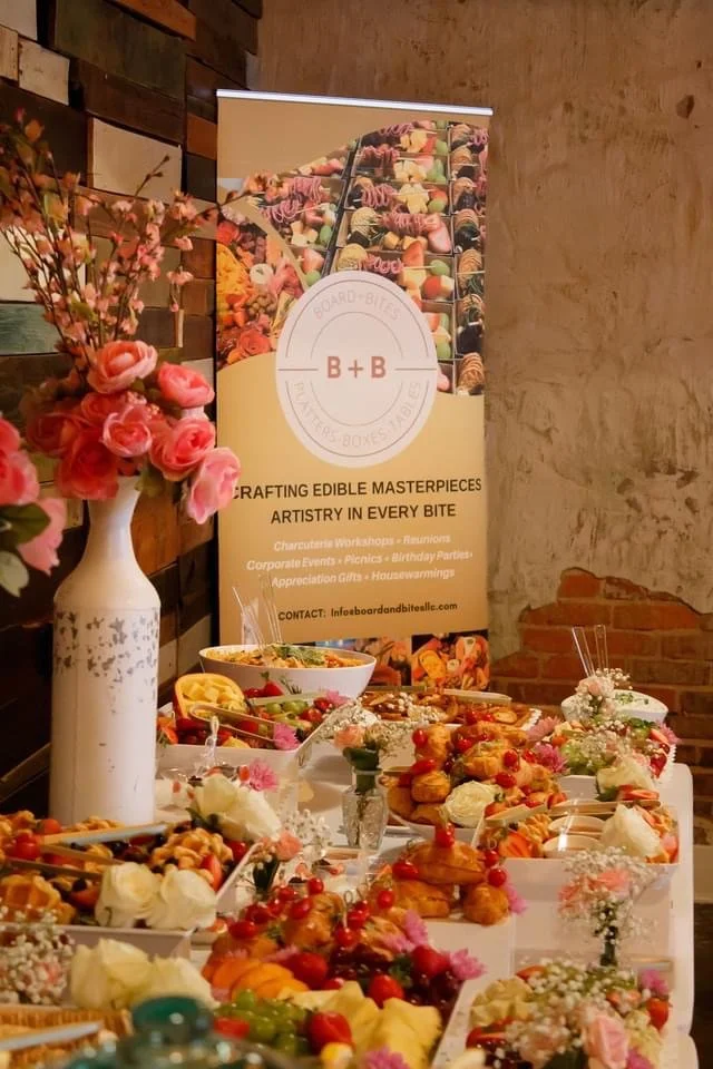 Table with a variety of colorful edibles and desserts, decorated with pink flowers and a banner from B + B catering company.