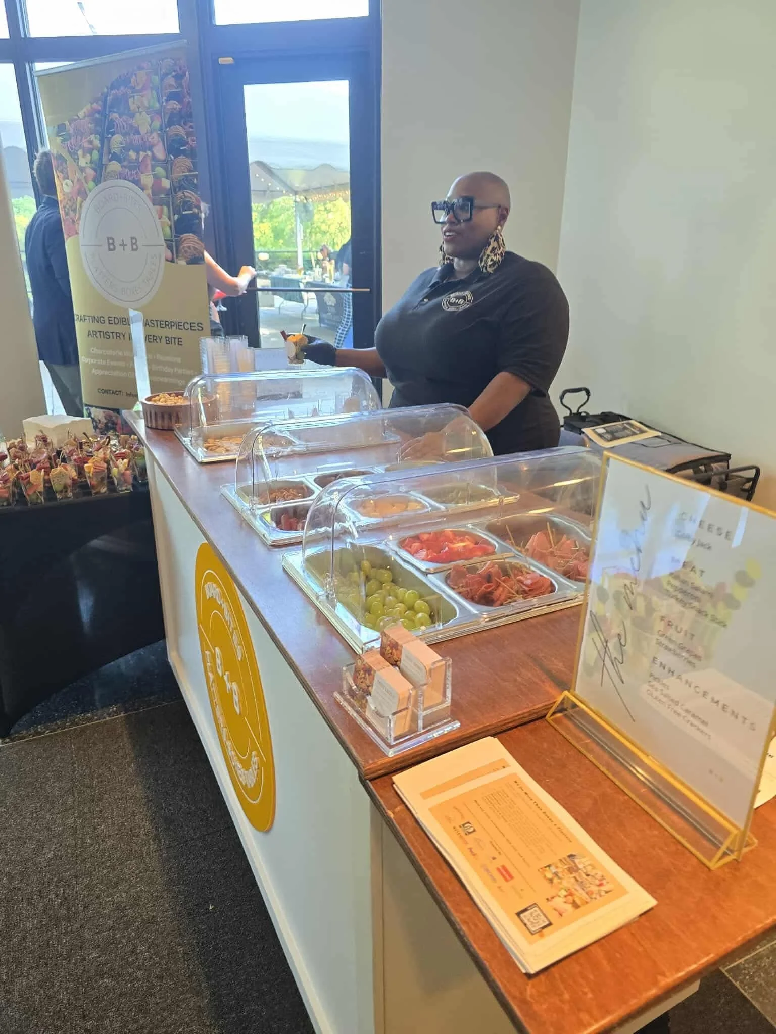 A woman with large earrings and sunglasses standing behind a buffet table with trays of cheese, grapes, and meats in a bright indoor setting.