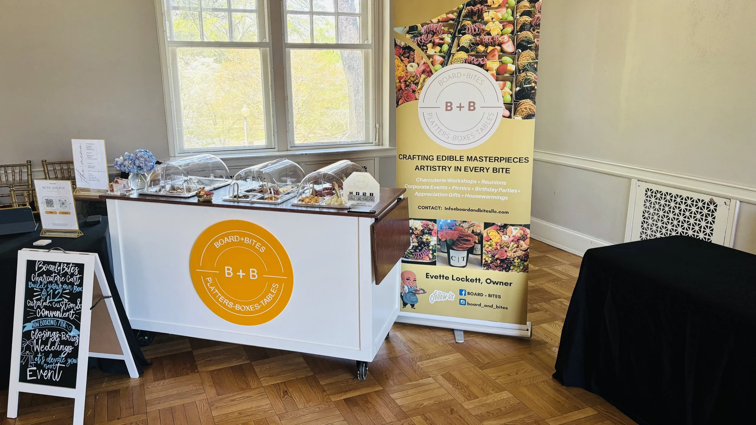 A buffet table with options under clear domed covers, a sandwich board with a black poster and white chalk-style lettering, and a standing yellow banner for Board+Bites, a food business specializing in edible arrangements and catering services. The r