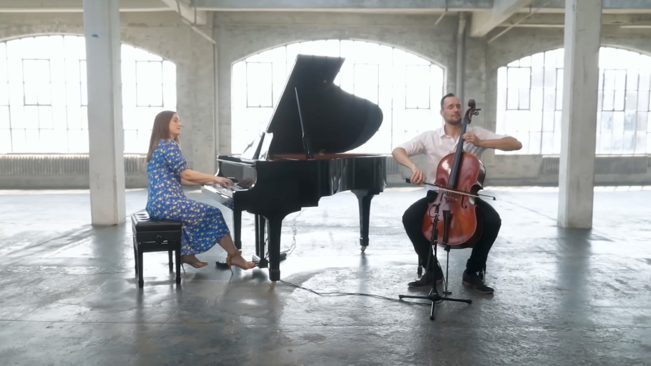 A woman playing a grand piano and a man playing a cello in a spacious, industrial-style room with large windows.