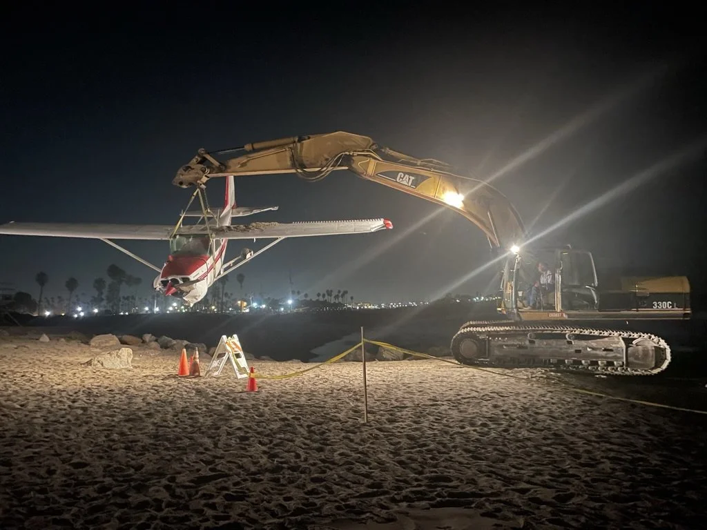 Nighttime scene of a small airplane and a large construction excavator working together on an airport runway. The excavator is lifting and positioning the airline for maintenance or repair. There are safety cones, a warning sign, and the area is cordoned off. City lights are visible in the distance.