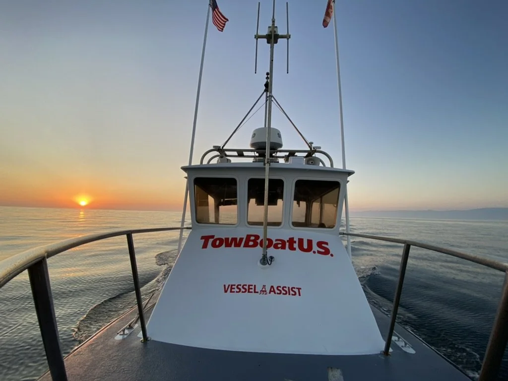 View from a boat at sunset showing the boat's bow, with the American flag, the company name 'TowBoatU.S.', and the words 'Vessel Assist'. The ocean is calm with a distant horizon and a colorful sky.