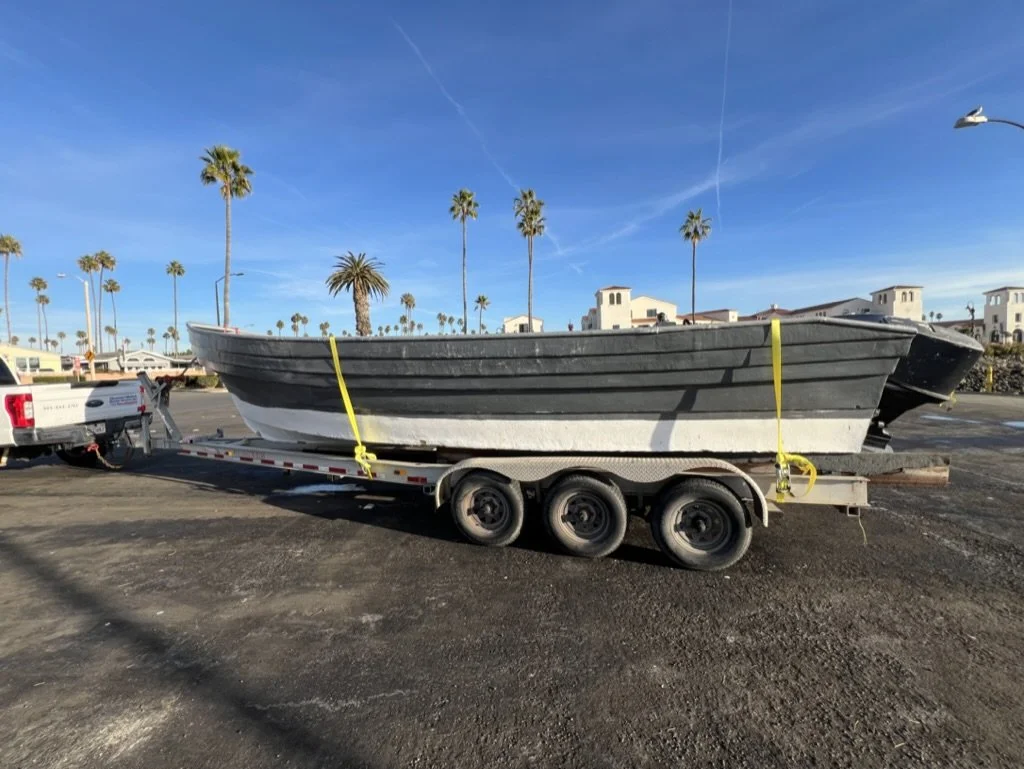 A boat on a trailer hitched to a pickup truck in a parking lot with palm trees and buildings in the background.