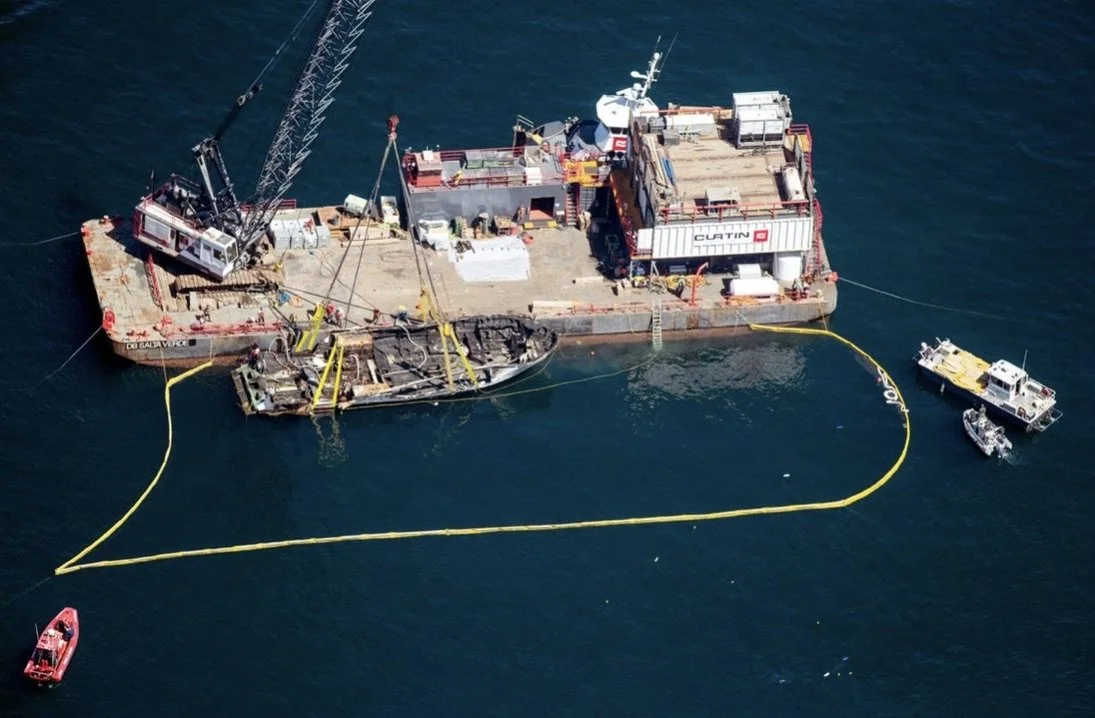 A sunken ship surrounded by yellow containment boom, with rescue boats nearby and a crane on a floating platform.