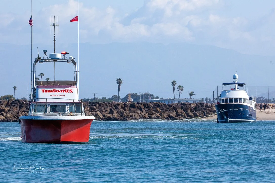 Two boats sailing in the water near a rocky shoreline with palm trees and mountains in the background.
