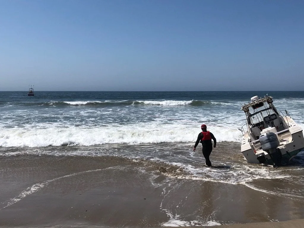 A person in a black wetsuit and red life jacket walking into the ocean near a tilted motorboat on the sandy beach, with a calm sea and clear blue sky.