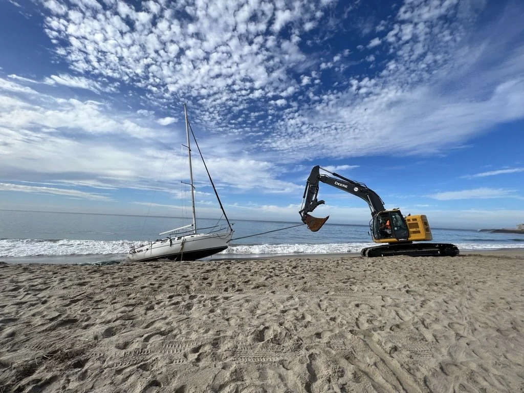 A construction excavator on a sandy beach pulling a small sailboat out of the water with a cloudy sky overhead.