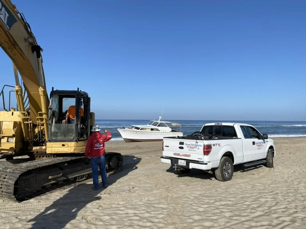 Construction equipment, a woman in a red jacket and white hard hat, a white boat on the beach, and a white pickup truck with vessel assistance logo near the ocean.