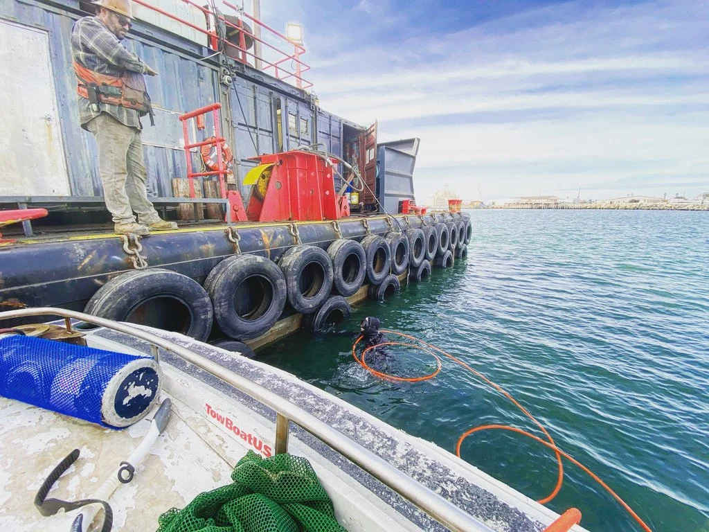 A worker stands on a dock near a large floating platform, observing a diver in the water with an orange safety line. The platform has large black tires along the edge and various equipment on top. The background shows a clear sky and industrial structures in the distance.