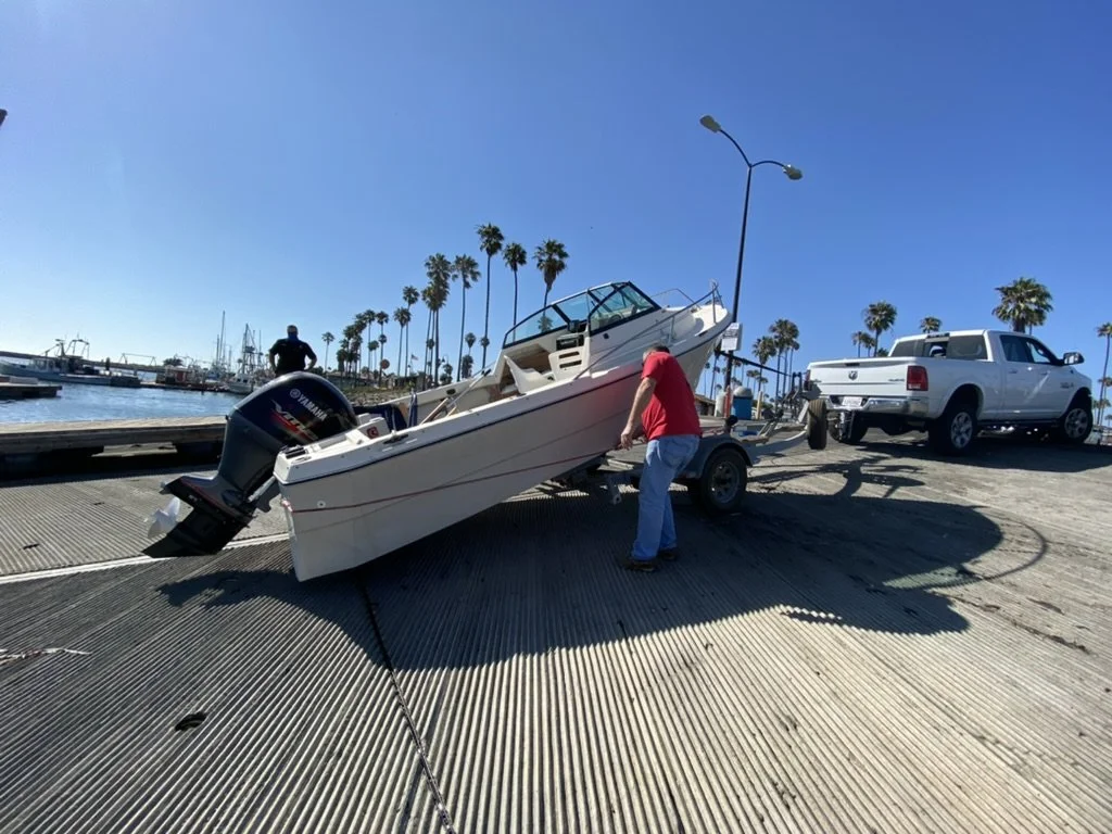People preparing a boat for launch at a marina with palm trees, a clear blue sky, and parked trucks.