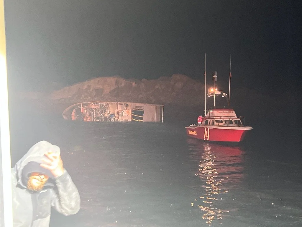 A person with a hooded jacket in the foreground, a flipped boat in the water, and a red rescue boat nearby at night.