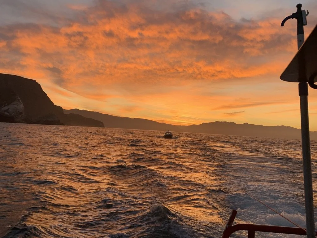 A sunset over a body of water with a boat in the distance, calm rippling waves, and distant mountains; part of a boat with a pole and a speaker on it in the foreground.