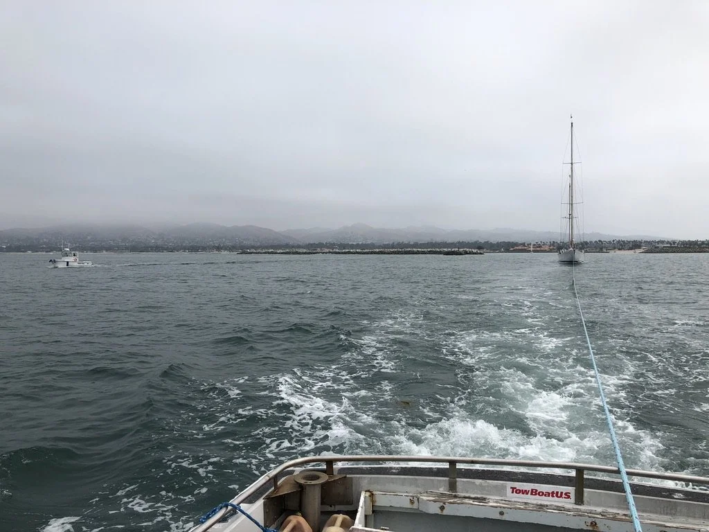 View from a boat showing the water, a small motorboat, and a sailboat in the distance under a cloudy sky with a city skyline and hills on the horizon.