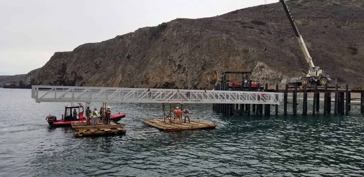 Construction workers and machinery installing a large metallic structure on stilts over water near a rocky shoreline.