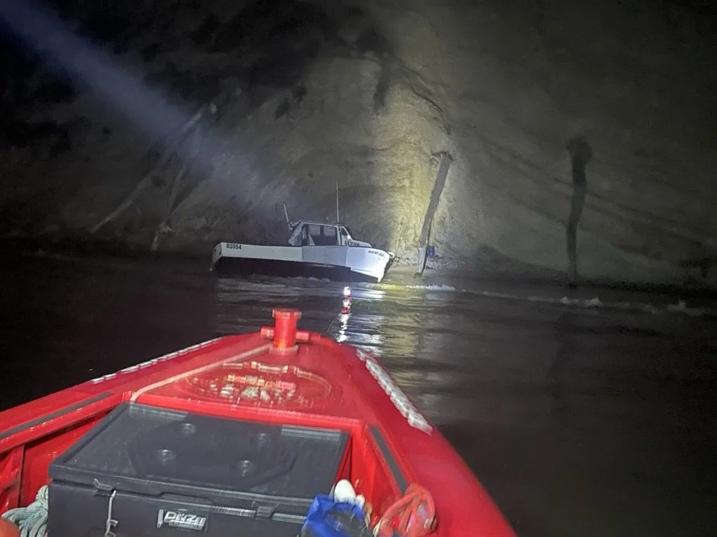 A boat with a red hull is approaching a submerged boat inside a dark, rocky cave or tunnel. The scene is illuminated by artificial light, revealing the rocky walls and water surface.