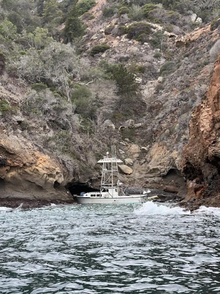 A white boat with a tall mast navigating through a narrow rocky water passage between steep, rugged cliffs covered with sparse trees and shrubs.