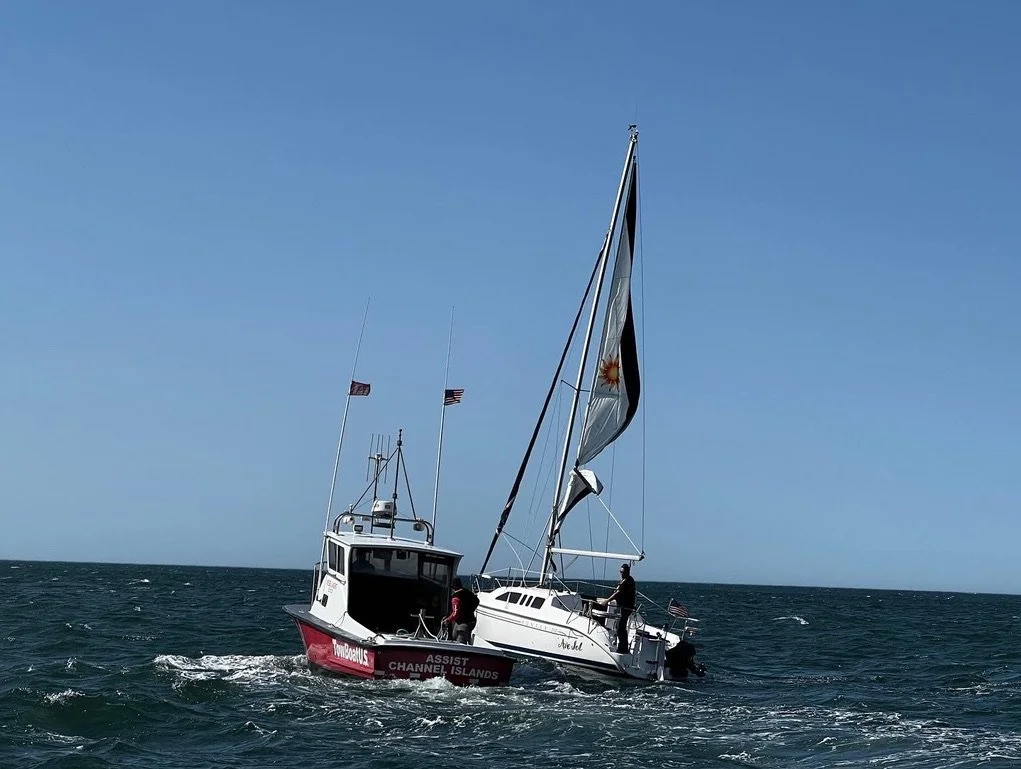 A sailboat capsized in the water with two people on it, flags flying, under a clear blue sky.