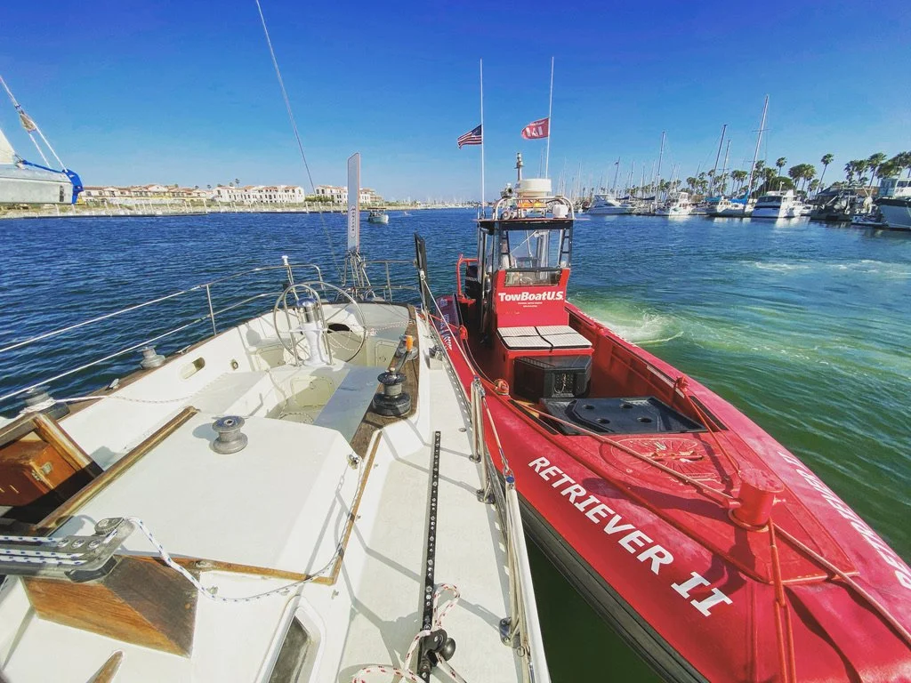 View from a yacht showing a red rescue boat named 'RETRIEVER II' docked nearby with a marina full of sailboats and yachts in the background on clear, sunny day.