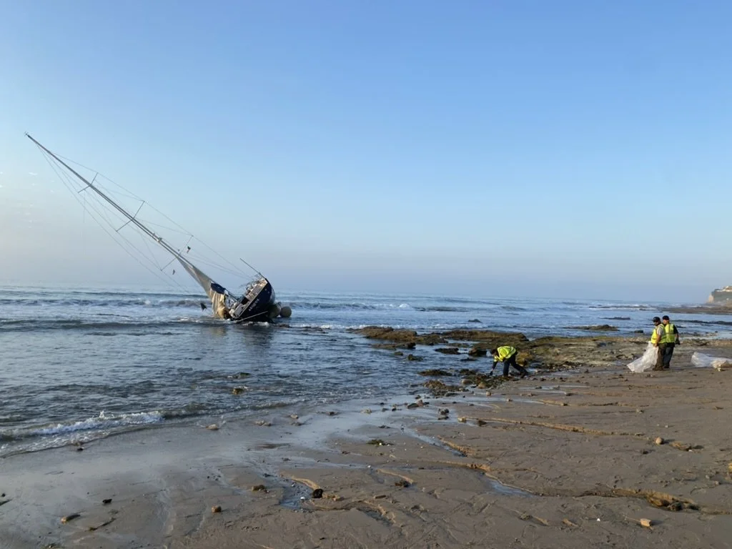 A sailboat tilted on its side in the shallow water near the shoreline, with three people in reflective vests working nearby.
