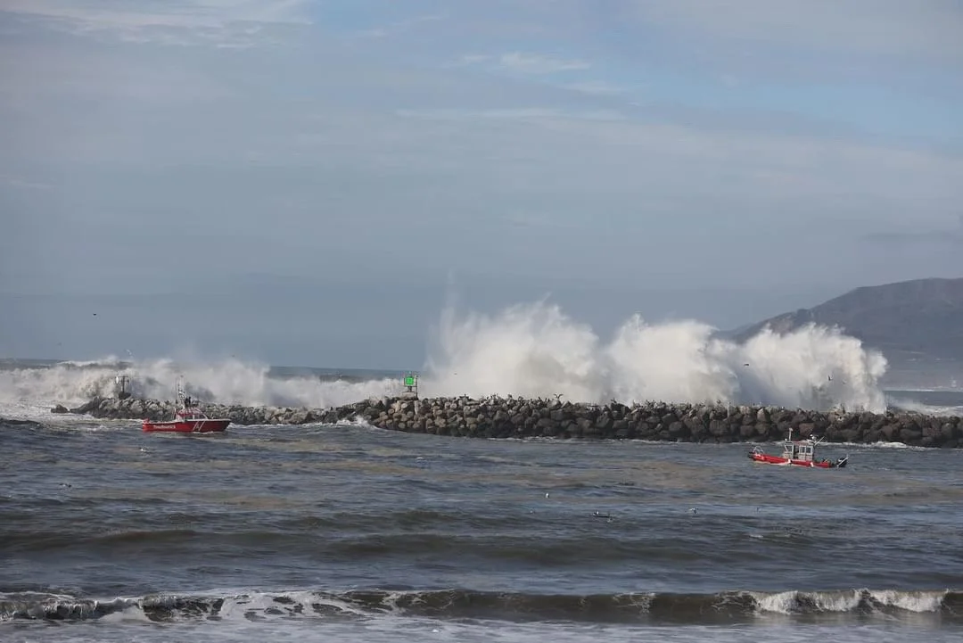 Waves crashing against a rocky breakwater with two red boats in the water nearby.