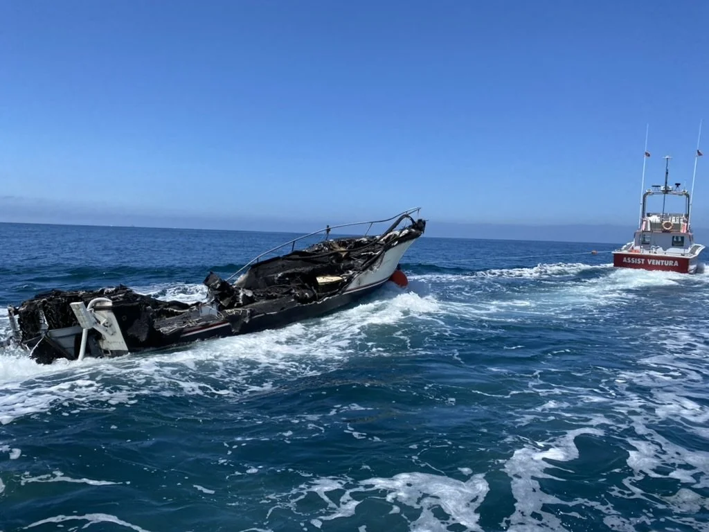 A damaged boat sinking in the ocean with a rescue boat approaching in the background.