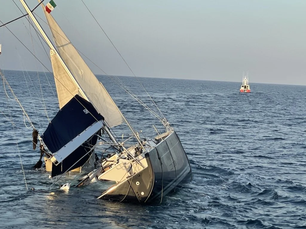 A sailboat capsized in the water with parts of the boat submerged, and another boat in the background on the open sea.