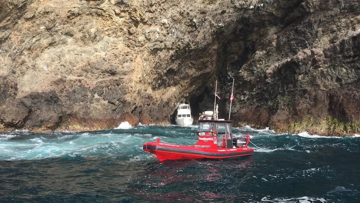 A red rescue boat near rocky cliffs with a cave entrance, and white boats in the water.