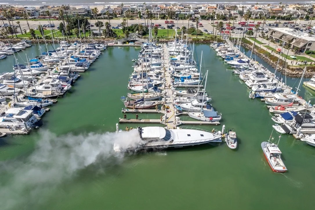 A marina with multiple sailboats and yachts docked, and a speedboat leaving a trail of smoke as it moves through the water.
