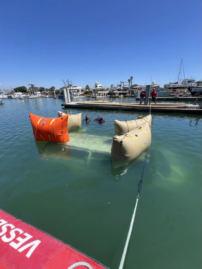 Two people in the water near a floating platform with large orange and beige inflatable buoys, with a marina and boats in the background under a clear blue sky.