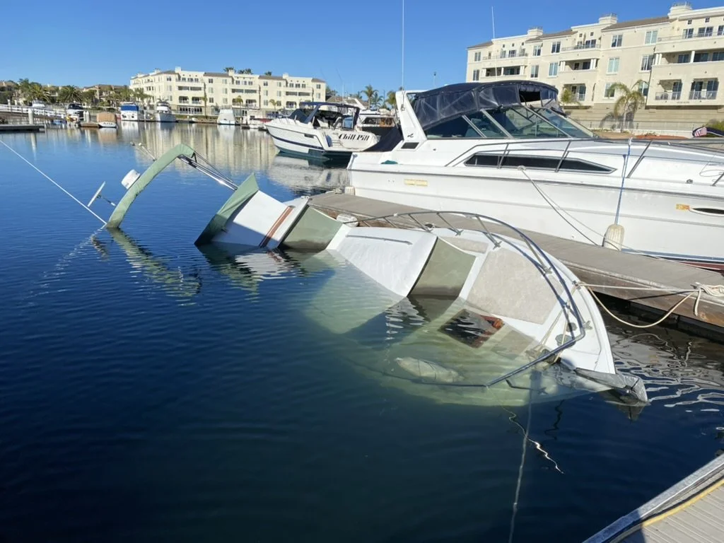 A partially submerged boat leaning to one side in the water at a marina, with luxury yachts and residential buildings in the background.