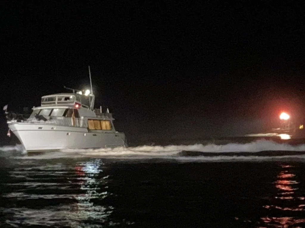 Nighttime photo of a white yacht on the water with a bright light, and another vessel in the distance with a red light, creating reflections on the water.