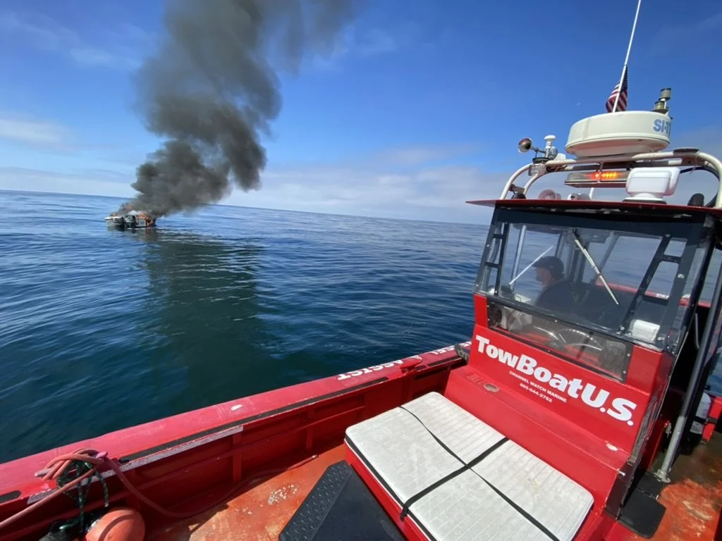 A fire on a boat in the water with black smoke billowing into the sky, viewed from a nearby patrol boat labeled 'TowBoatU.S.'