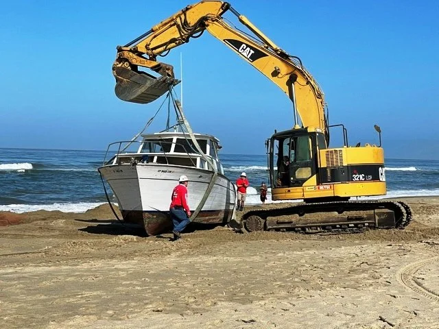 Construction equipment lowering a boat onto a beach with workers nearby