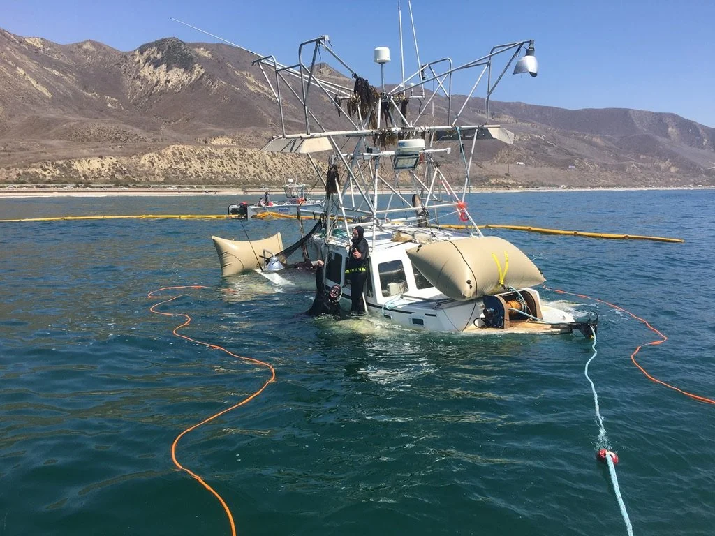 A partially submerged boat with a large metal structure on top, floating in the water near a shoreline with mountains in the background. Crew members are working on the boat, surrounded by floating barriers and equipment.