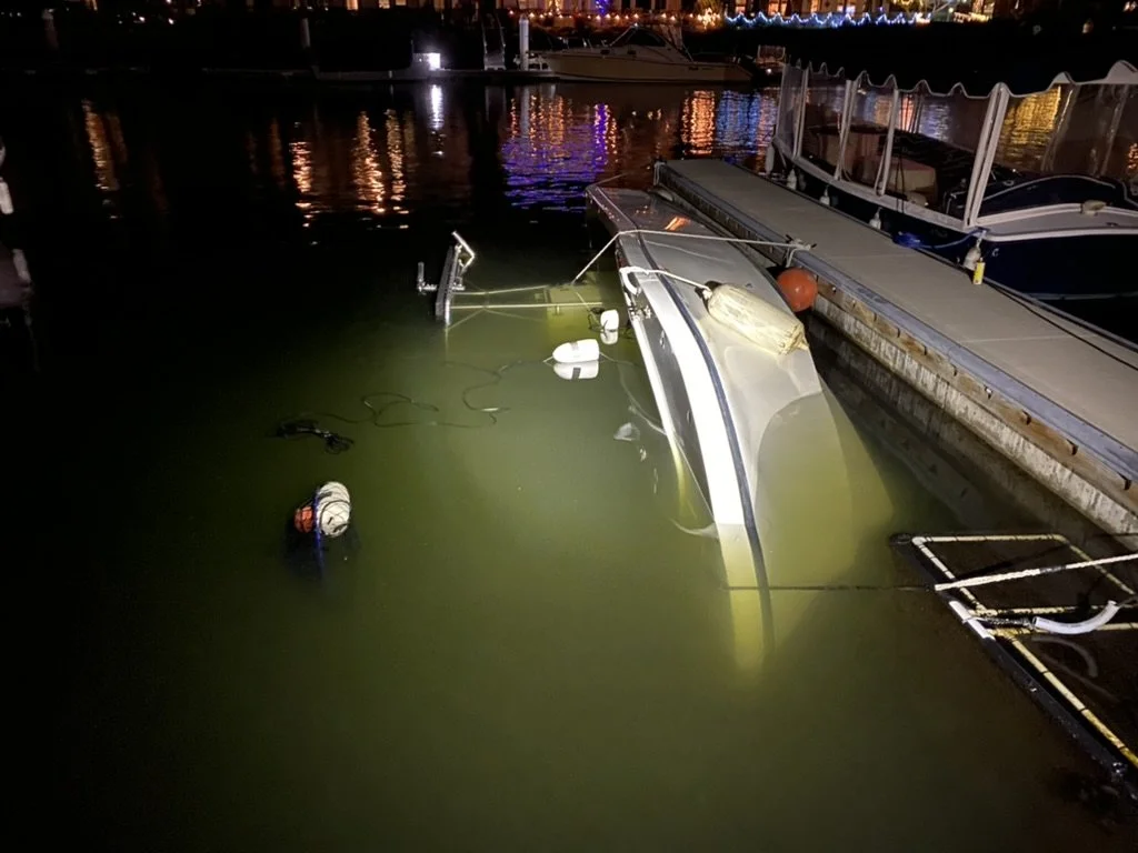 A boat is capsized and partially submerged in murky water next to a dock at night, with lights reflecting on the water's surface.