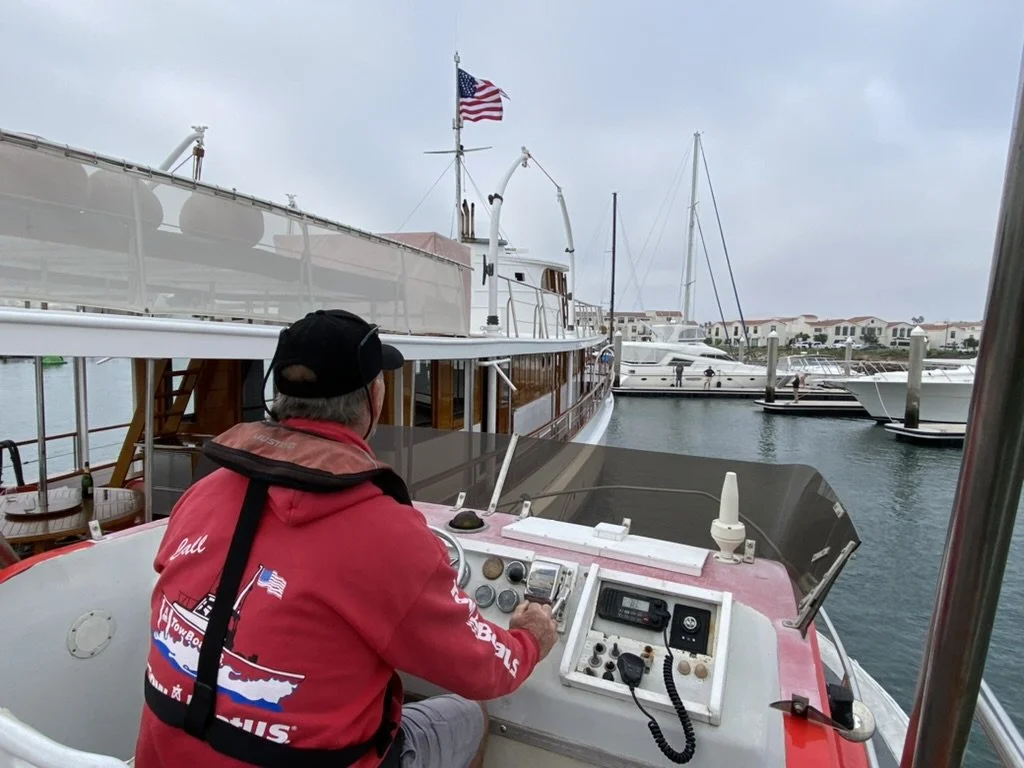 A man in a red jacket and black cap operating a boat's control panel in a marina with multiple yachts and sailboats docked.