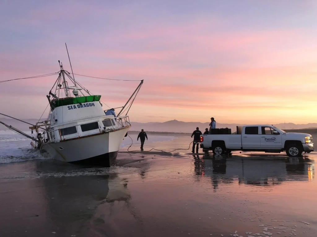 A boat named 'Sea Dragon' is beached on the shore during sunset; three men are working nearby with a pickup truck.