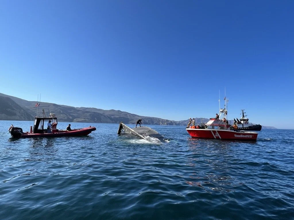 Three boats on the ocean with two people on each boat and two people on the sinking boat, while a whale is visible near the boats, with a rocky shoreline and hilly landscape under a clear blue sky in the background.