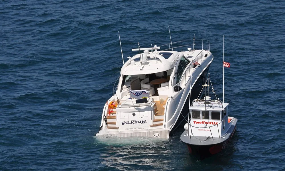 An aerial view of a white luxury yacht named Valkyrie sailing on the ocean, with a smaller vessel labeled TowBoatUS alongside it.