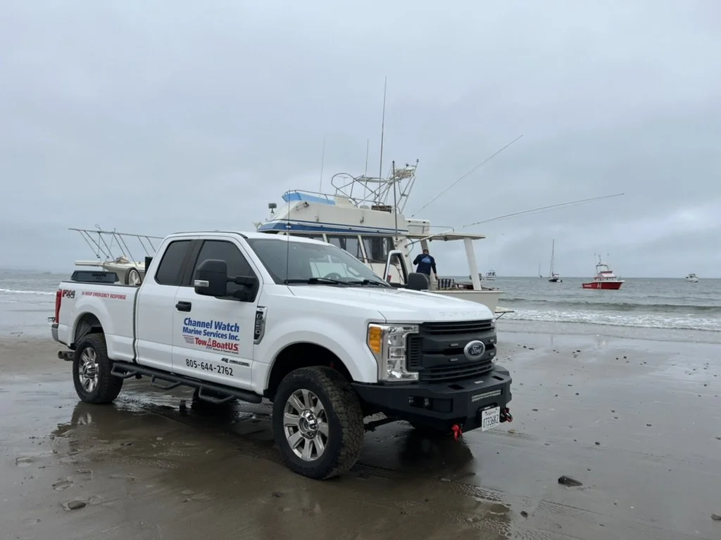 A white truck parked on a beach with a boat and water in the background.
