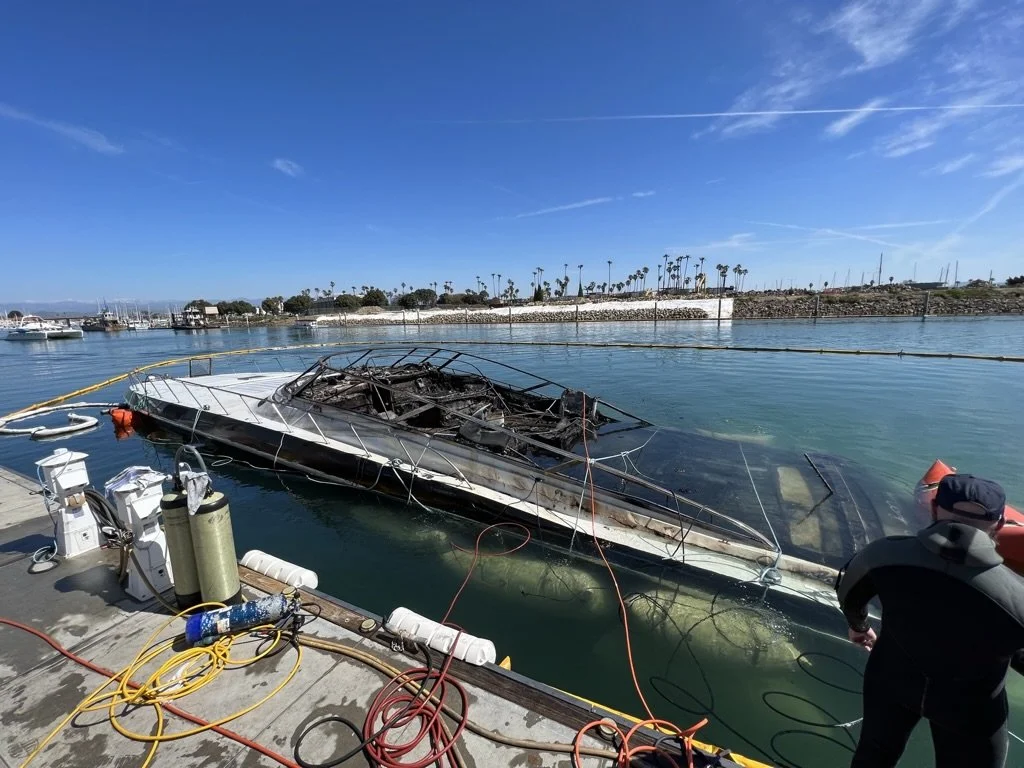 Burned and damaged boat docked at a marina with electrical equipment nearby and a man observing the scene.
