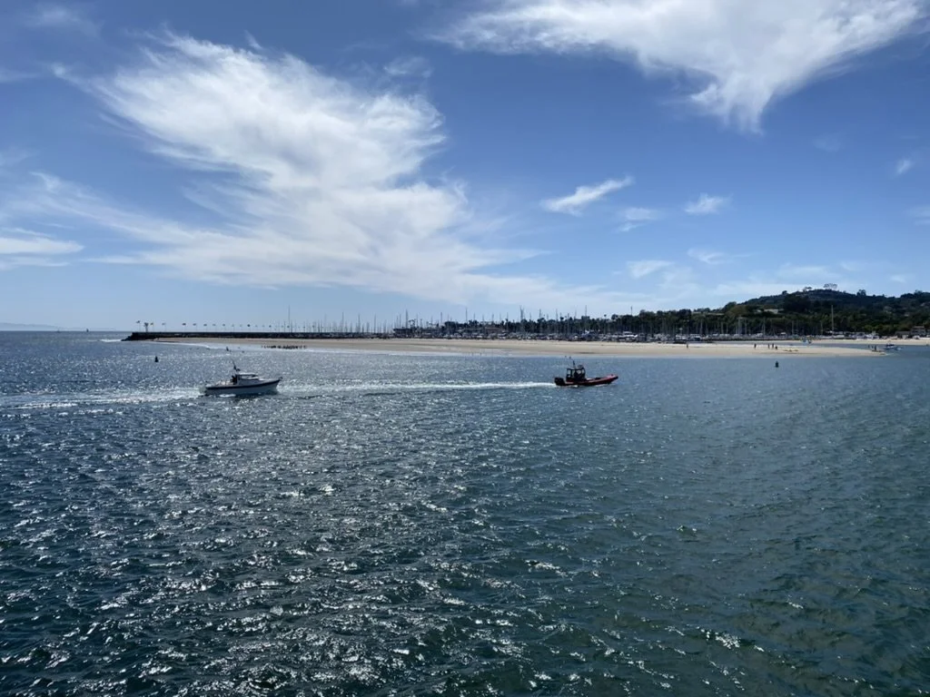 Two boats sailing on calm ocean waters near a sandy beach with a marina and hillside in the background under partly cloudy skies.