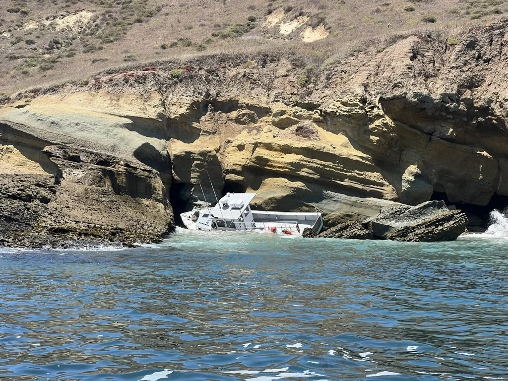 A boat partially crashed into a rocky coastal area with cliffs and large boulders, surrounded by blue water.