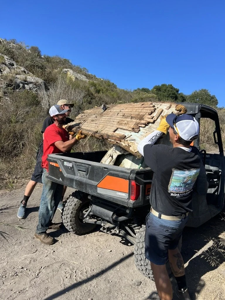 Three men are removing a damaged, old wooden roof from a small vehicle with a cargo bed on a dirt trail in a hilly, rocky area as part of a cleanup or repair project.
