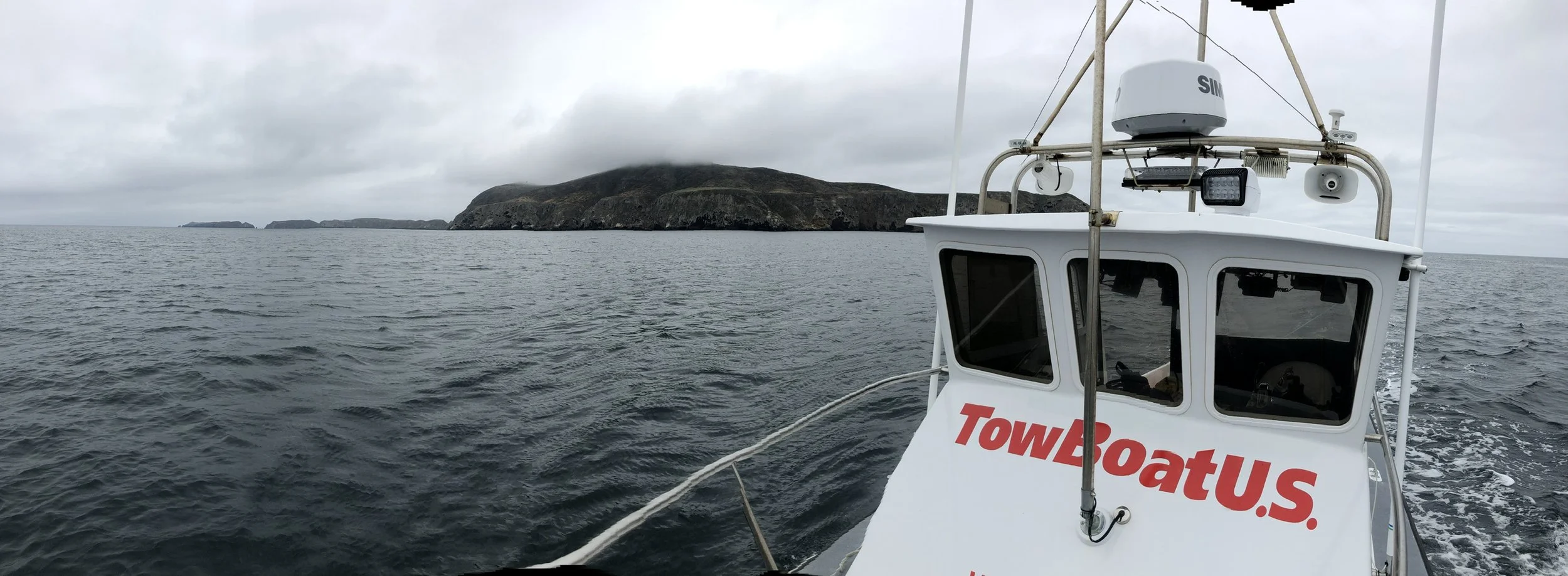 View from a boat named TowBoatU.S. on open water with a mountainous island in the background. The boat's cabin has various navigation and communication equipment, and the sky is overcast.